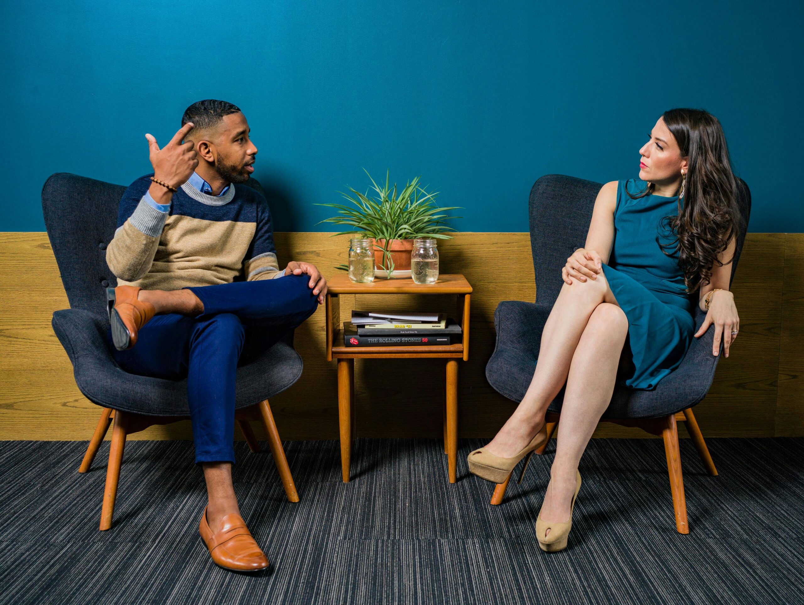 A woman in a teal dress sitting while speaking with a man.